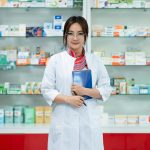 Female pharmacist in a lab coat standing confidently in a well-organized pharmacy.