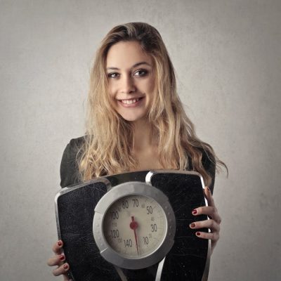 Young woman with blond hair smiling, holding a vintage bathroom scale indoors.