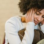A woman with curly hair sits indoors, appearing stressed and holding her head in her hands.